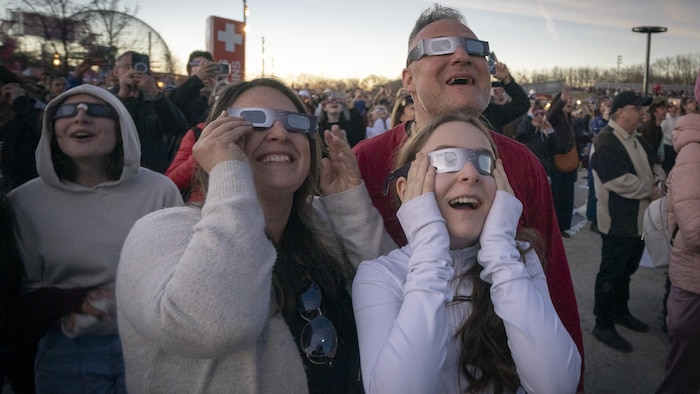 Deux adultes et deux jeunes qui portent des lunettes protectrices pour les éclipses sourient, éblouis, en regardant vers le ciel.