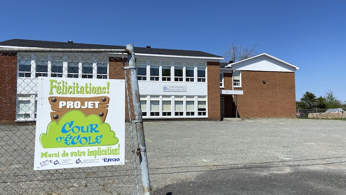 Photo extérieure d'une école, un bâtiment de deux étages. À l'avant-plan, une affiche de félicitations pour un projet de cour d'école accrochée sur une clôture.