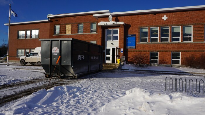 La façade d'une école avec un conteneur à déchets et des fenêtres placardées.