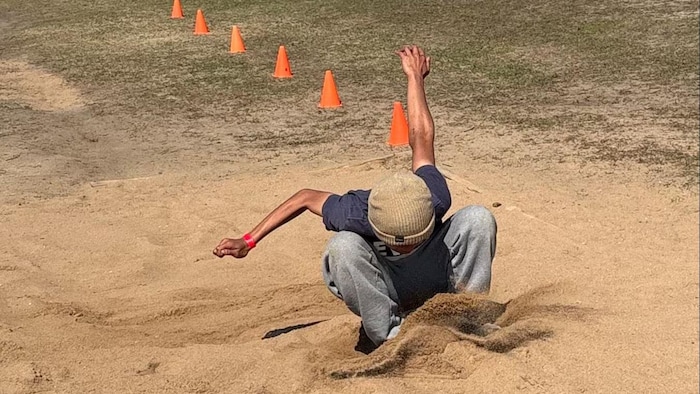 Un jeune athlète en pleine extension vient d'atterrir dans la fosse de sable, capturé à l'instant précis de l'impact lors d'une compétition de saut en longueur.