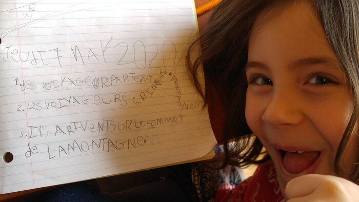 Une fillette au large sourire montre une courte rédaction dans un cahier ligné.