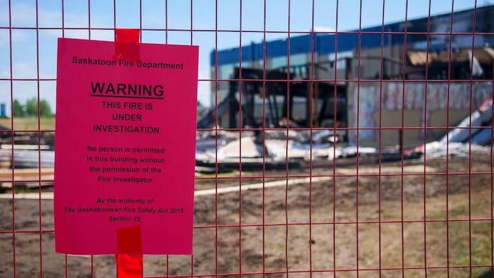 Un papier indiquant que le Service d'incendie de Saskatoon mène une enquête. En arrière-plan, les restes d'un édifice qui a été la proie des flammes.