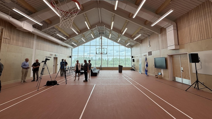 Un gymnase d'école, un panier de basketball à l'avant-plan, et quelques personnes au loin.