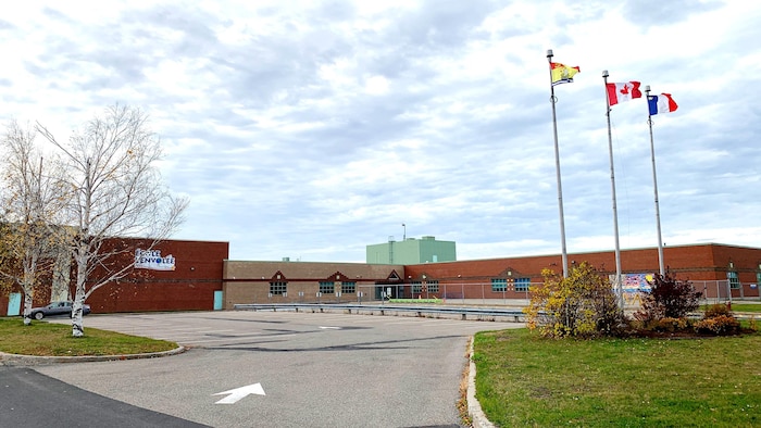 Bâtiment de l'école et 3 mâts où flottent les drapeaux de l'Acadie, du Nouveau-Brunswick et du Canada.