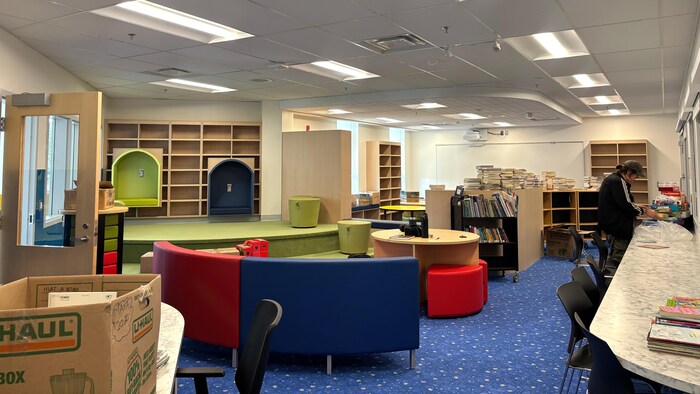 Une bibliothèque scolaire dans laquelle on voit une boîte de déménagement en carton posée sur une table, et une personne au loin qui trie des livres.