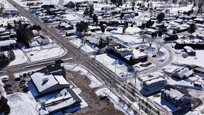 Cette photographie aérienne présente un village nordique sous un manteau neigeux. Une route principale traverse le quartier résidentiel composé de maisons individuelles et de petits commerces. En arrière-plan, une forêt dense borde les limites de la zone urbanisée.