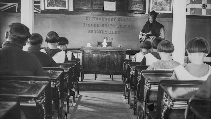 Des élèves, garçons et filles, sont assis à des pupitres (de dos) devant une religieuse qui tient un livre ouvert à l'avant de la classe.