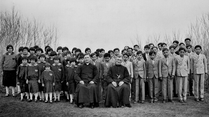 Des élèves de l'École Brocket, en Alberta, en 1930, sont debout en uniformes d'élèves derrière deux religieux assis.