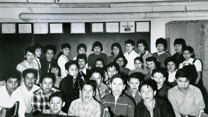 Des élèves, garçons et filles, posent pour la photo dans une classe. Ils sont debout, derrière une rangée de pupitres, et des dessins (motifs autochtones?) sont accrochés au mur derrière le groupe.