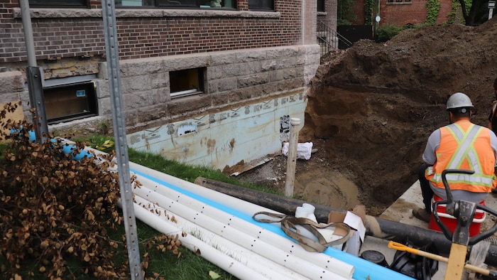 Un travailleur s'affaire près d'un chantier. Un trou est bordé d'un large tas de terre et des tuyaux sont empilés tout près. Une partie du solage de l'école est à découvert.
