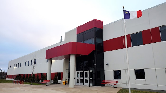Enseigne de l'école Mosaïque devant un bâtiment et un drapeau de l'Acadie qui flotte sur un mât.
