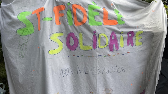En diverses couleurs, les mots « St-Fidèle solidaire, non à l'expulsion » sont écrits, dessinés de mains d'enfants, sur un grand tissu. Le message est accroché avec des pinces sur un arbuste.