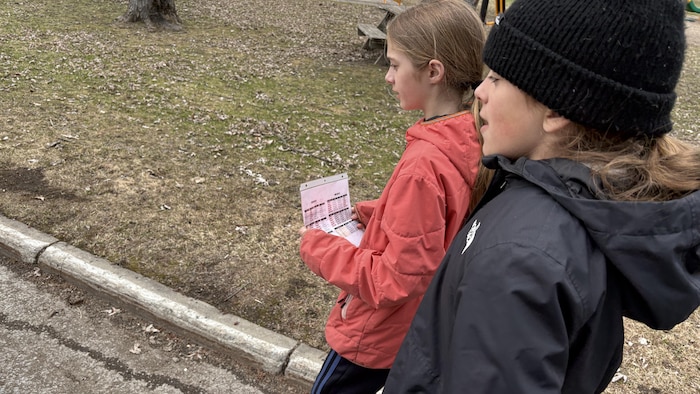 Deux enfants marchent à l'extérieur près d'un parc. L'une porte un manteau rose et tient une feuille avec des tables de calculs, tandis que l'autre porte un manteau et une tuque noirs.