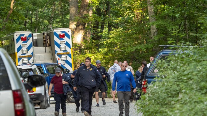 Un groupe d'hommes marche dans une forêt près de véhicules stationnés des deux côtés du chemin.