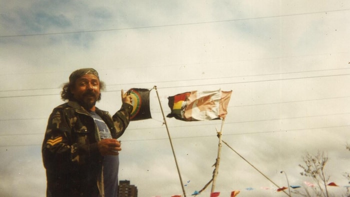Une photo d'un homme relativement jeune, coiffé d'un bonnet et vêtu d'une veste militaire, qui est debout et pointe des drapeaux qui flottent au vent.