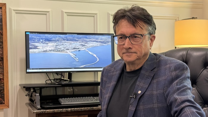 L'élu assis à son bureau. Derrière lui se trouve un écran d'ordinateur sur lequel se trouve une photo du port de Matane.