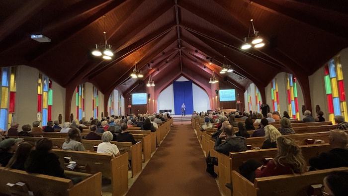 Plan large sur une église avec des paroissiens assis sur des bancs qui regardent une conférencière sur scène.