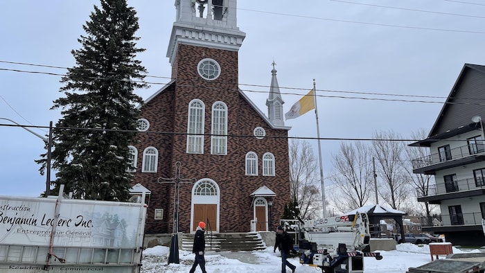 La remorque d'un entrepreneur général en construction est stationnée devant l'église, ainsi qu'un chariot élévateur. Deux travailleurs sont à l'œuvre.