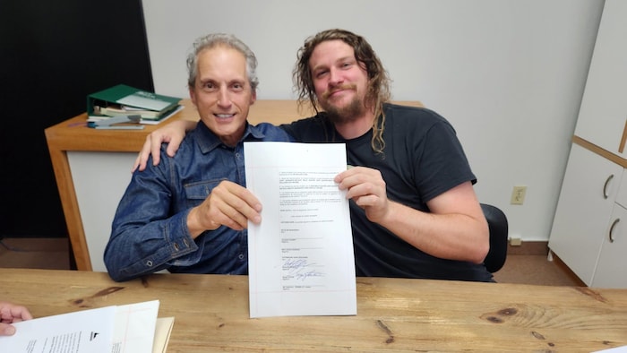 Deux hommes souriants tiennent un document portant plusieurs signatures devant l'objectif.