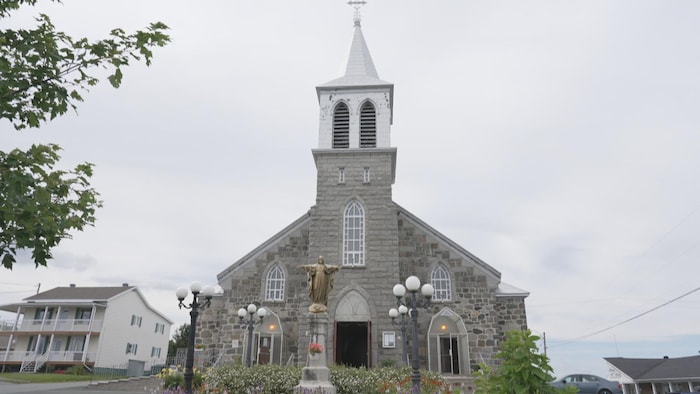 Une église. Devant, un parterre de fleurs blanche et une statue de Jésus en croix.