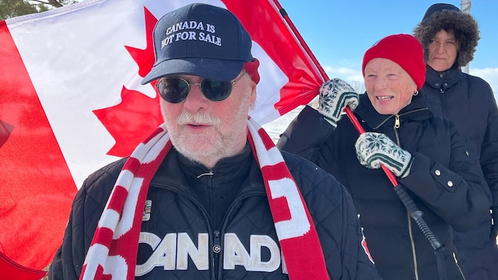 Un homme en premier plan portant des vêtements en soutien au Canada avec deux femmes derrière un drapeau canadien.