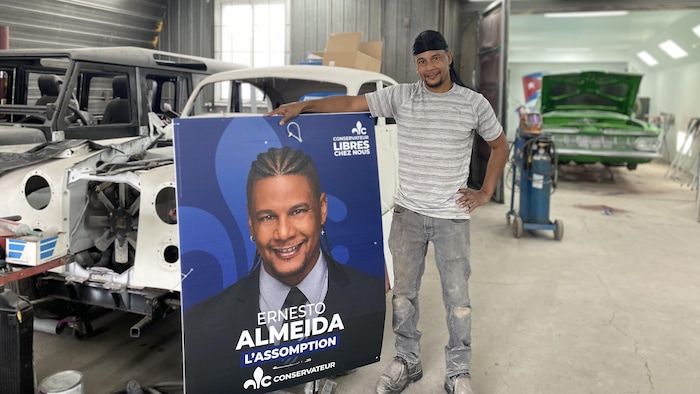 Un homme pose avec une pancarte électorale à son effigie devant des voitures en réparation dans un garage.