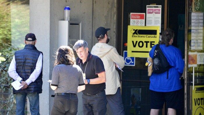 Des gens font la file pour voter. Un employé, de face, parle à une femme. En arrière-plan, une affiche indique « Vote ».