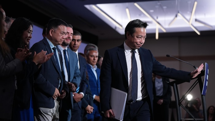 Ken Sim, l'air déçu, quitte le lutrin devant les applaudissements de son équipe. 