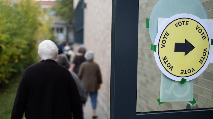 Gros plan sur un autocollant « VOTE » avec une flèche sur une vitre, devant des gens vus de dos.