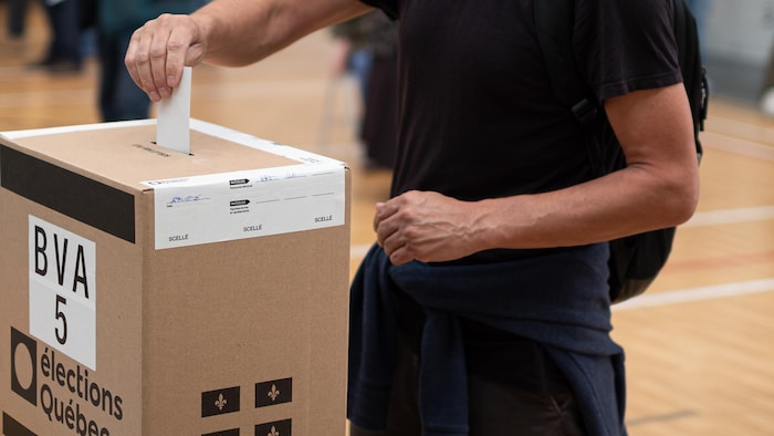 Une personne portant un sac à dos dépose son bulletin dans une urne en carton d'Élections Québec.