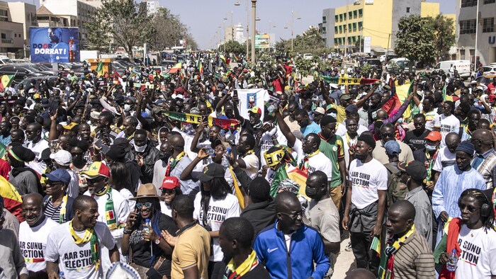 Beaucoup de personnes manifestent dans la rue en tenant des pancartes et en portant des foulards aux couleurs du drapeau du Sénégal. 