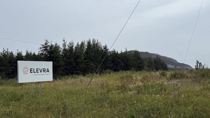 Une affiche indique Elevra Lithium Amérique du Nord à l'entrée d'un chemin. On voit une montagne de roches à l'arrière.