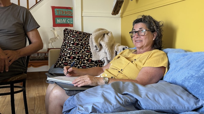 Une femme portant des lunettes est assise avec un carnet de notes et un stylo à la main, le regard tourné vers le groupe.