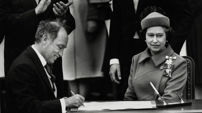 Photo d'archive sur laquelle on voit la reine Elisabeth 2 assise en compagnie de Pierre Elliott Trudeau qui signe proclamation constitutionnelle.