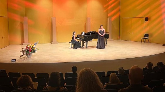 Les deux femmes en robe noire sont sur scène, dans une salle de concert, et donnent un récital devant un auditoire.