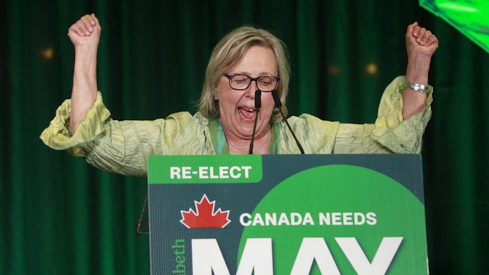 Elizabeth May parle au micro et lève les bras en signe de victoire à la tribune le soir des élections.