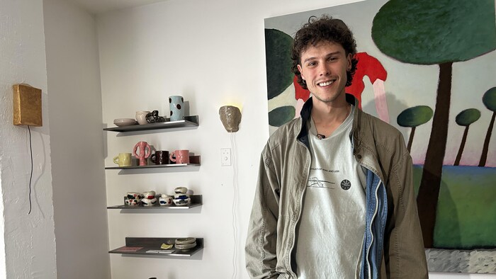 Un jeune homme souriant, debout dans une boutique moderne. Il porte une veste kaki sur un t-shirt gris clair et un col en jean, avec les mains dans les poches. À sa gauche, des étagères flottantes présentent des céramiques d'art et des publications de design.