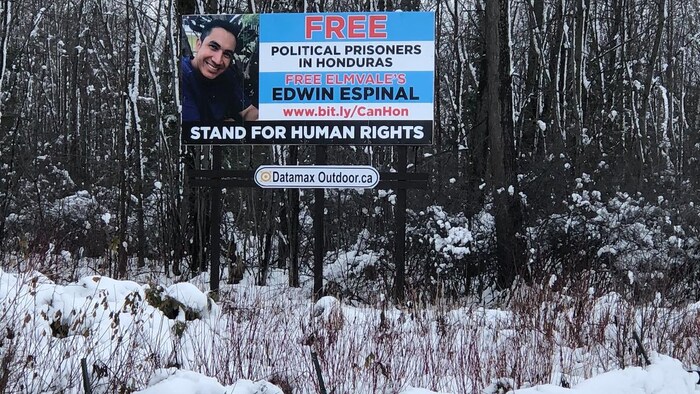 Un cartel publicitario frente a un bosque cubierto de nieve muestra la imagen de Edwin Espinal con las palabras "Free Political Prisoners in Honduras - Free Elmvale's Edwin Espinal - Stand for Human Rights"