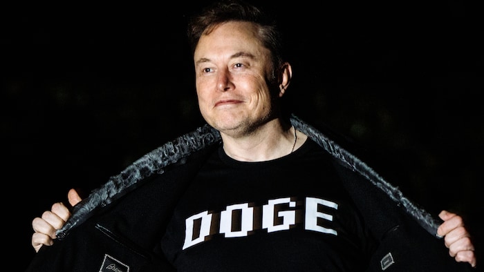 Elon Musk en train d'ouvrir son veston pour découvrir un tee-shirt sur lequel est écrit "DOGE".