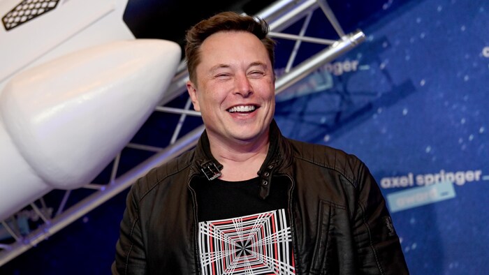 Elon Musk, souriant, avec une fusée derrière lui.