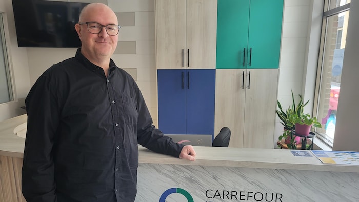 Un homme portant des lunettes et une chemise noire pose derrière un comptoir de réception. Sur le devant du meuble, on lit l'inscription « Carrefour Jeunesse-Emploi Saguenay ». L'arrière-plan montre des armoires colorées et une plante verte.