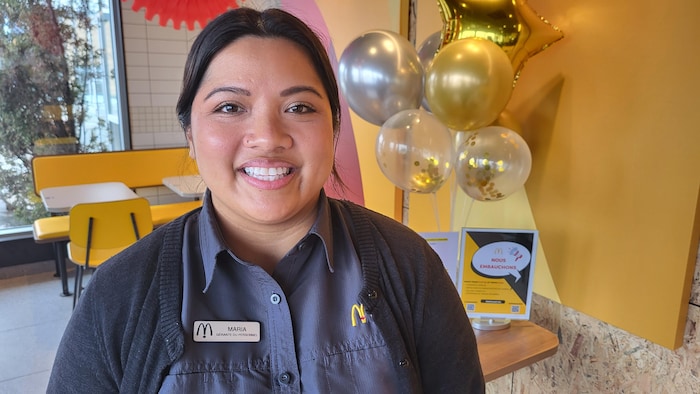 Maria, une employée souriante de McDonald's en uniforme, pose devant des décorations festives composées de ballons dorés et argentés. À côté d'elle, un petit présentoir indique « Nous embauchons », créant une ambiance accueillante à l'intérieur du restaurant.
