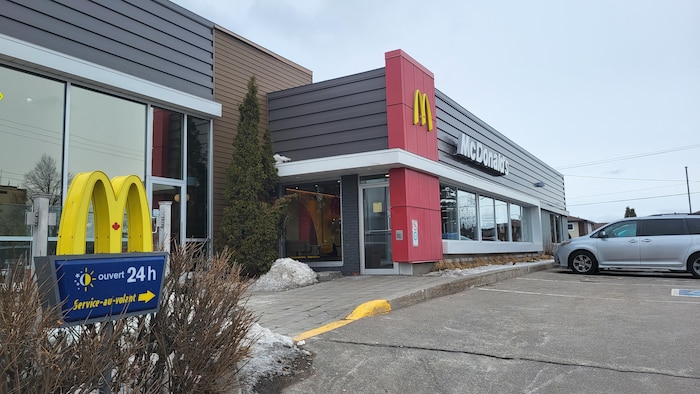 Cette photo montre l'extérieur d'un restaurant McDonald's au Canada par temps gris. On aperçoit une enseigne indiquant « ouvert 24 h », un service au volant, une voiture stationnée et des restes de neige sur les bordures du terrain.