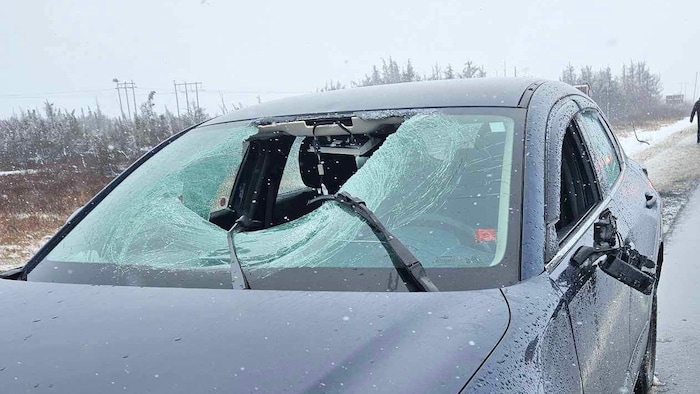 Un grand trou au milieu du pare-brise défoncé d'une voiture sur le bord de l'autoroute sous la neige.