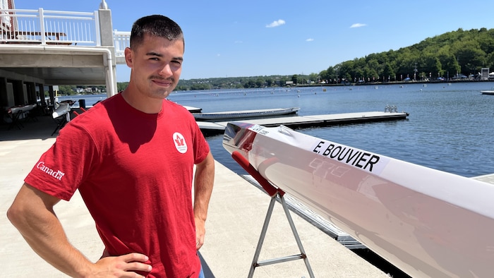 Un athlète en chandail rouge d'Équipe Canada est au bord du lac, les mains sur les hanches, à côté d'un canoë blanc sur lequel son nom est écrit.