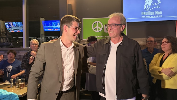 Deux hommes se tiennent par les épaules en souriant dans une salle pleine de gens.