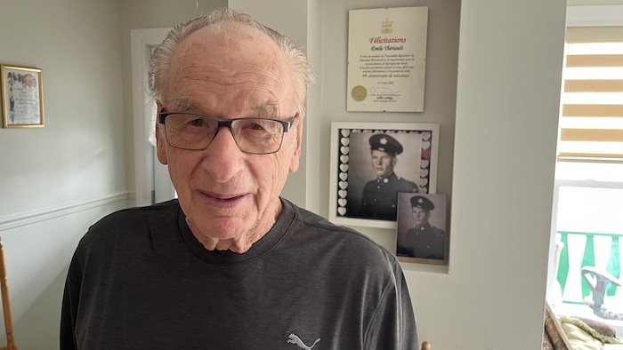 Homme âgé avec lunettes posant devant une photo d'un militaire.