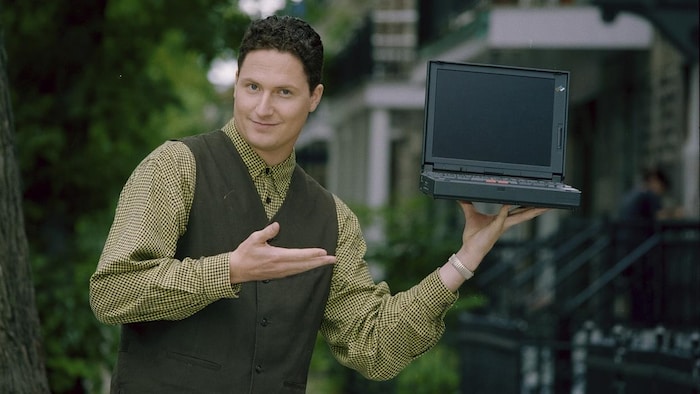 Jean-Hugues Roy qui présente un ordinateur portable qu'il tient d'une main.