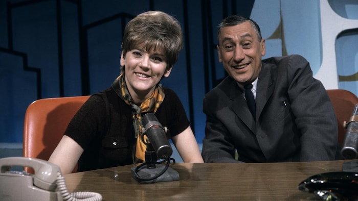 Dans un studio de télévision, les animateurs Clémence Desrochers et Roger Baulu, souriants, sont assis derrière un pupitre où reposent un téléphone et un micro de table pour l'émission du 9 mai 1968. 