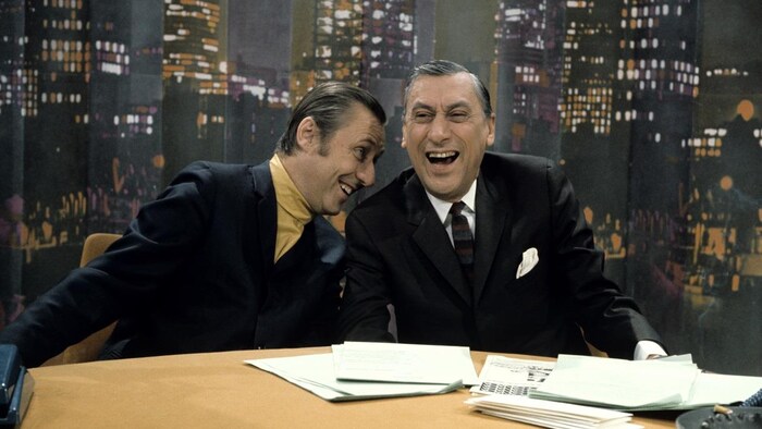 Dans un studio de télévision, les animateurs Jacques Normand et Roger Baulu discutent, assis derrière leur pupitre à l'émission du 28 mars 1969.